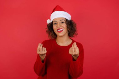 Noel şapkası takan ve kırmızı süveter giyen kıvırcık saçlı genç bir kadının kırmızı duvar arkasında poz veren bir stüdyo resmi.  