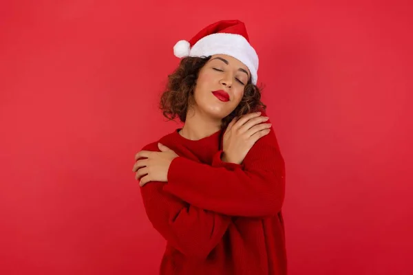 穿着圣诞帽和红色毛衣的漂亮女人在鲜红的背景下拥抱自己的画像 — 图库照片