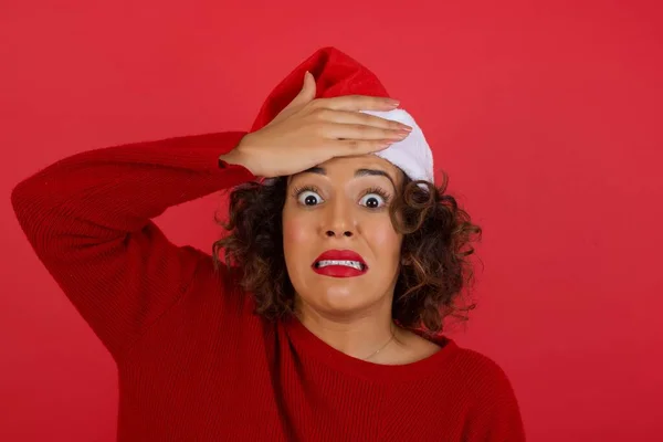 美丽的黑发女人 头戴圣诞帽 身穿红色毛衣 头戴红色背景 手挽手 脸上带着恐惧和遗憾的表情 — 图库照片