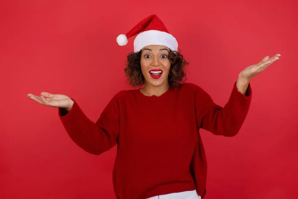 头戴圣诞帽 头戴红色毛衣的快乐女人做了个受欢迎的手势 她的胳膊在红色的背景上举过头顶 — 图库照片