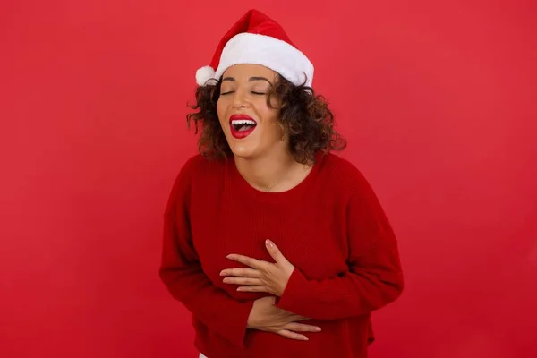 头戴圣诞礼帽 身穿红色毛衣的快乐女人两手交叉 笑着讲笑话 穿着休闲装和圆形眼镜 站在屋里 — 图库照片