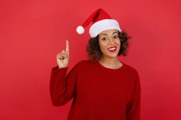 穿着圣诞帽和红色毛衣的年轻貌美女子的服装肖像有聪明的表情 举起一只手指 想起自己不要忘了告诉重要的事 室内环境 — 图库照片