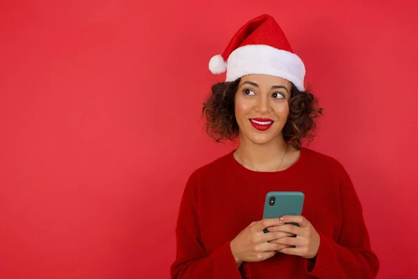 一个戴着圣诞帽子 头戴红色毛衣的年轻貌美女子在明亮的红色背景下 用手机拍照的照片 — 图库照片