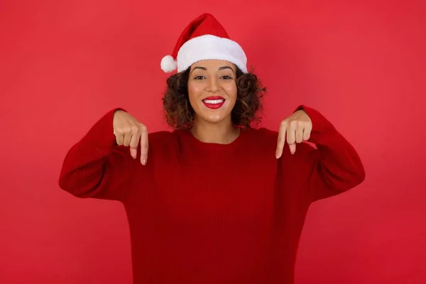 クリスマスの帽子と赤いセーターを身に着けている自信と自信に満ちたカリスマ的な女性は 幅広く微笑み 赤い壁の上に体を指差す役割を望んでいるように自分自身を促進します — ストック写真