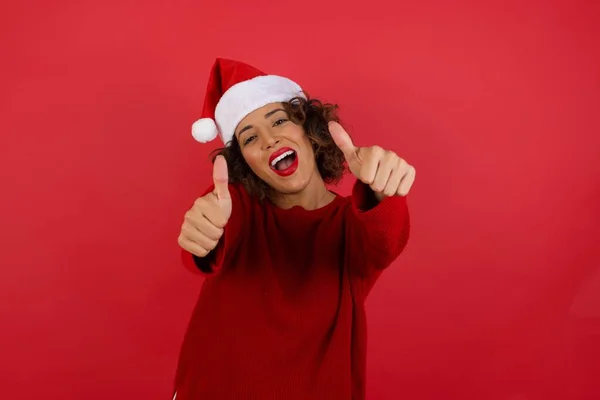 年轻女人戴着圣诞帽 红色背景的红色毛衣 双手做着积极的手势 微笑着竖起大拇指 为成功感到高兴 看着镜头 — 图库照片