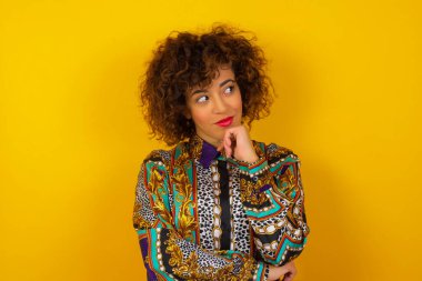 Hayalperest Arap kadın renkli gömlek giyiyor, hoş bir ifade takınıyor, günlük kıyafetler giyiyor, yan bakıyor, elini çenesinin altında tutuyor, hoş bir şeyler düşünüyor, arka planda poz veriyor..