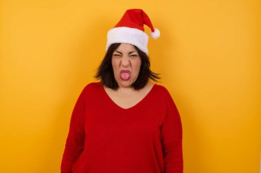 Stresli, orta yaşlı, Noel şapkası takan, gri duvara karşı poz veren, suratını asan, bir şeyden tiksindiğini gösteren Arap bir kadın..