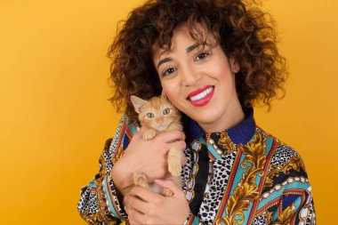 Elinde kedi olan beyaz bir kadın sarı duvara yaslanmış, mutlu görünüyor. 