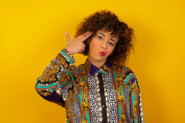 Renkli gömlek giyen mutsuz genç Arap kadın silah atışı taklidi yapıyor intihar jesti yapıyor iki parmağını şakakların üzerinde tutuyor günlük kıyafetler giymiş şaşkın bir ifade içeride duruyor..