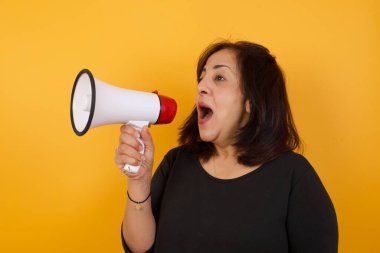 Orta yaşlı bir kadın elinde megafonla sarı arka planda duruyor.