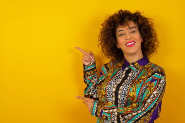 Reklam konsepti. Pozitif, güzel, renkli tişörtlü, ışıl ışıl gülümseyen Arap kadın işaret parmağıyla işaret ediyor ve boş fotokopi odasına bakıyor..