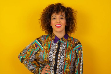 Renkli tişört giyen neşeli Arap kadının stüdyo fotoğrafı elini kalçasında tutuyor, geniş bir şekilde gülümsüyor, doğum gününde arkadaşlarının hazırladığı sürprizi bekliyor, arka planda izole edilmiş..