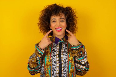 Güçlü, sağlıklı, beyaz dişlere sahip olma kavramı. Arkaplanda izole edilmiş, kusursuz beyaz dişlerini gösteren, ışıl ışıl gülümseyen renkli gömlekli Arap kadının yakın portresi..