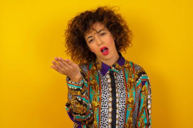 What the hell are you talking about, nonsense. Studio shot of frustrated Arab woman wearing colorful shirt    gesturing with raised palm, frowning, being displeased and confused with dumb question over wall.