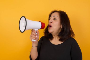 Orta yaşlı bir kadın elinde megafonla sarı arka planda duruyor.