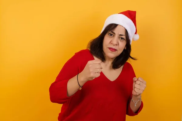 Portrait of attractive middle aged woman wearing Christmas hat with trendy hairstyle and clothes holding hands in boxing position going to fight.