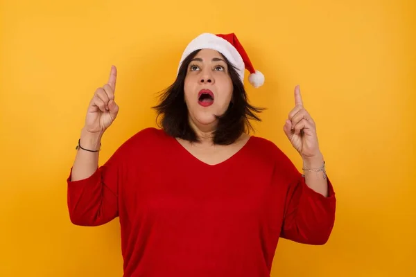 迷人的中年妇女头戴积极向上的圣诞礼帽 用食指指尖尖尖着 穿着休闲装 笑容满面 看那里 — 图库照片