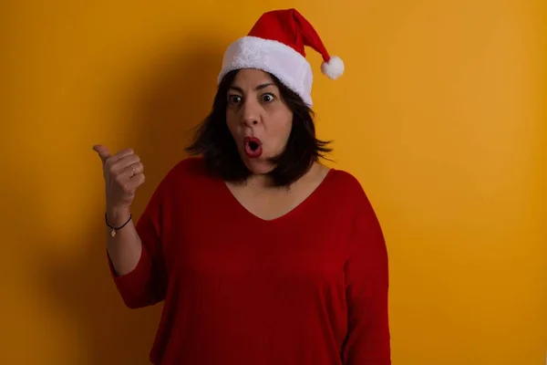 Horizontal shot of  shocked arab middle-aged woman wearing Christmas hat points with thumb away, indicates something on blank wall over background. Check this out.