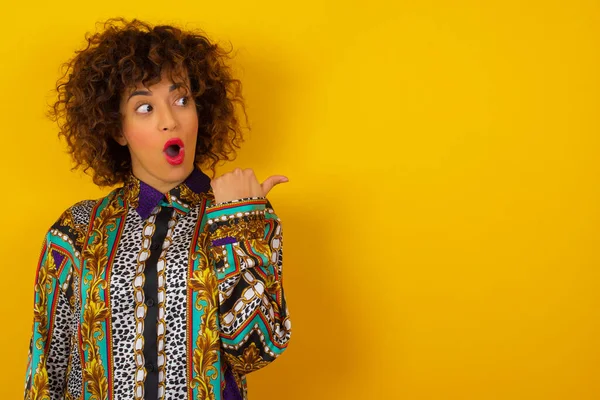 Horizontal shot of pretty young Arab woman wearing colorful shirt   points with thumb away, indicates something on blank wall isolated over background. Check this out. Ethnicity concept.