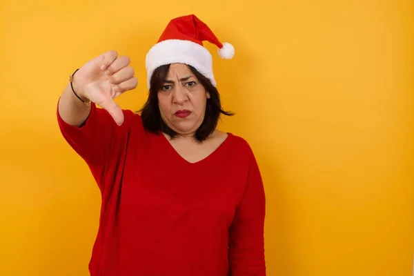 Discontent arab middle-aged woman wearing Christmas hat shows disapproval sign, keeps thumb down, expresses dislike, frowns face in discontent.