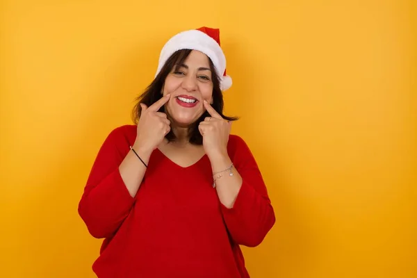 Close up portrait of happy middle aged woman wearing Christmas hat with beaming smile pointing on her perfect clear white teeth