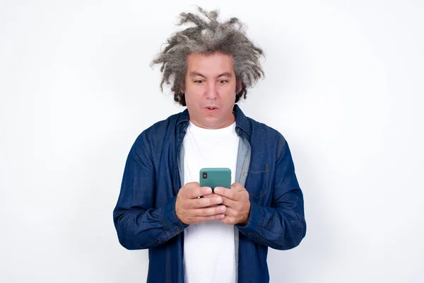白い壁の背景に隔離されたアフロ灰色の髪を持つハンサムな男は バグの目で見て 現代のスマートフォンを保持し 友人からの予期しないメッセージを受信し リマインダーを読む — ストック写真