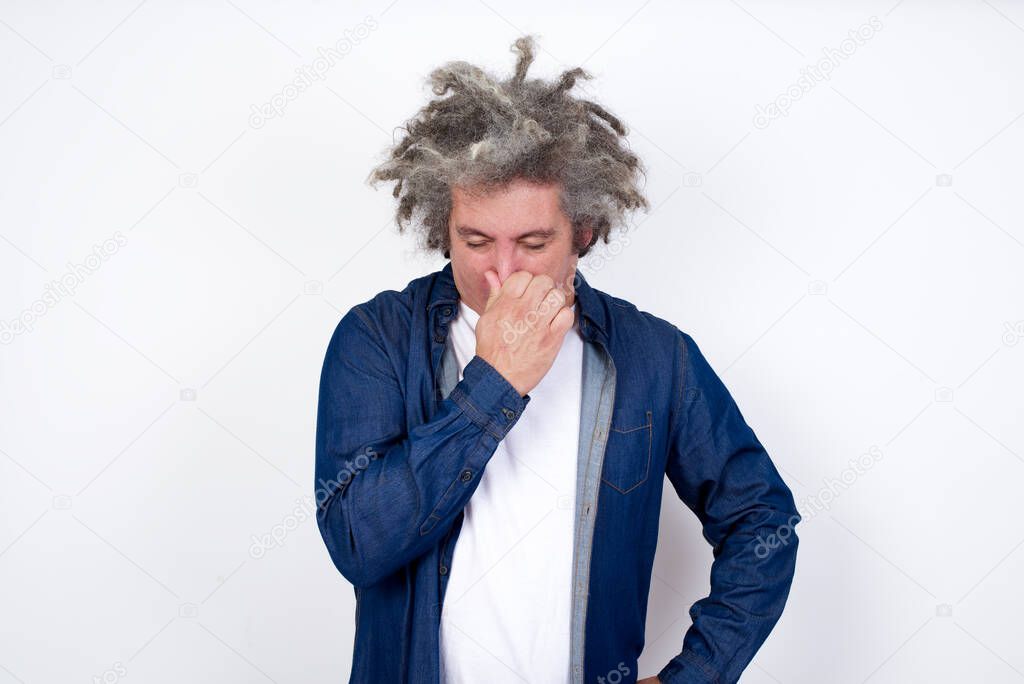 Handsome man with afro gray hair standing against white background, holding his nose because of a bad smell.