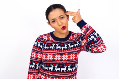 Mutsuz, genç, güzel bir kadın Noel süveterini beyaz duvara dayayıp, silah atışı taklidi yapıyor intihar jesti yapıyor. Parmağını şakaklarda tutuyor..