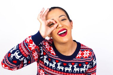 Genç, güzel bir kadın, beyaz duvarda Noel süveteri giyiyor, mutlu bir yüzü var, gülümsüyor..