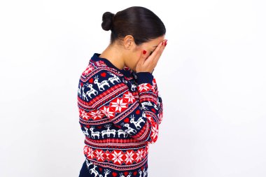 Üzgün, genç, güzel bir Arap kadın Noel süveteri giyip beyaz duvara yaslanmış elleriyle yüzünü kapatıyor ve ağlıyor..