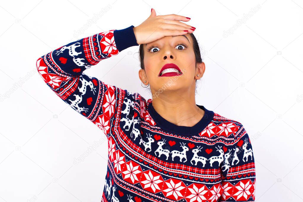 Oops, what did I do? Young beautiful  woman wearing Christmas sweater against white wall, holding hand on forehead with frightened and regret expression. 