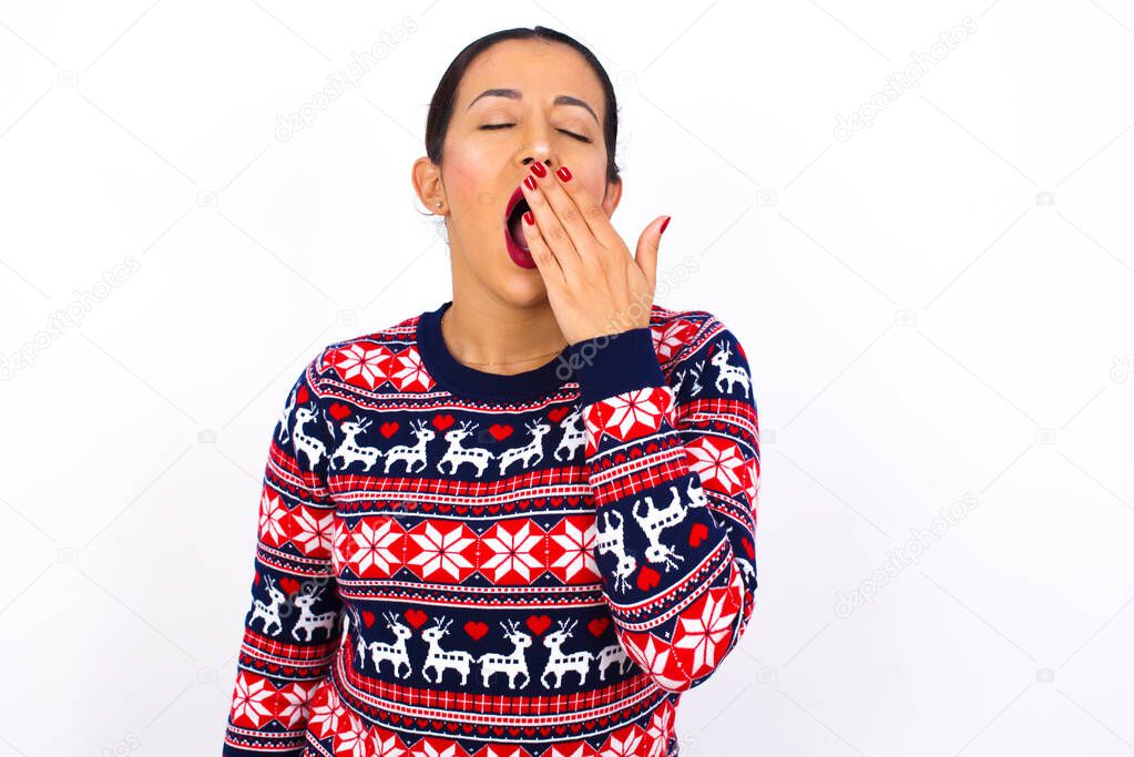 Sleepy  Young beautiful Arab woman wearing Christmas sweater yawning with messy hair, feeling tired after sleepless night, yawning, covering mouth with palm.