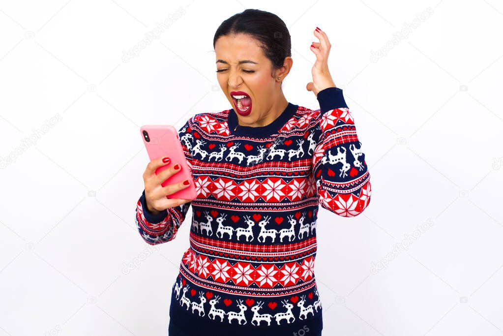 Photo of outraged annoyed  Young beautiful Arab woman wearing Christmas sweater holds cell phone, makes call, argues with colleague,  expresses negative emotions. People and anger.