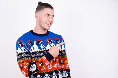 Mutlu, güler yüzlü beyaz adam Noel süveteri giyiyor beyaz duvara bakıyor ve eliyle işaret ediyor. Alan ve reklam konseptini kopyala.