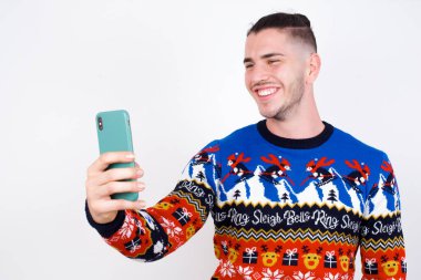 Isolated shot of pleased cheerful Young handsome Caucasian man wearing Christmas sweater against white wall,, makes selfie with mobile phone. People, technology and leisure concept clipart