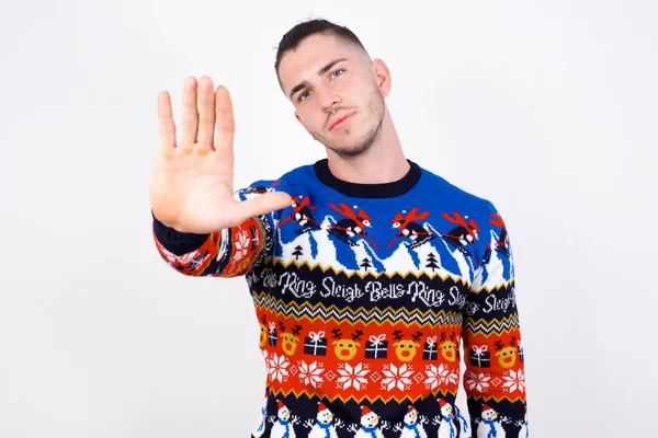 白い壁にクリスマスセーターを着て手のひらで停止ジェスチャーをする白人男性 顔にネガティブで深刻なジェスチャーで警告表現 — ストック写真
