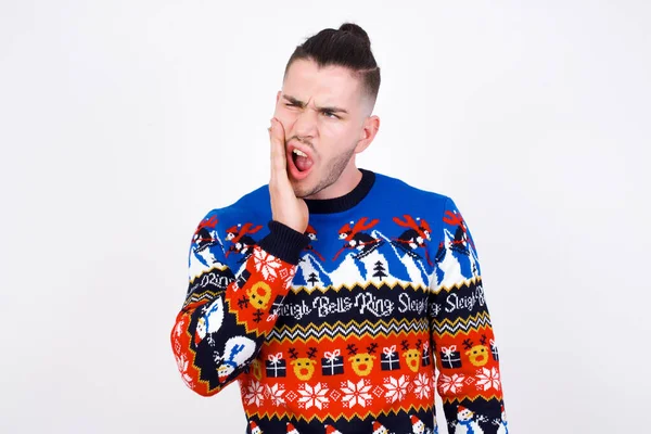 歯磨きコンセプト 白い壁の感じの痛みに対してクリスマスのセーターを身に着けている若い白人男性の屋内撮影 手で頬を握り 痛みを伴う表情でカメラを見ている — ストック写真
