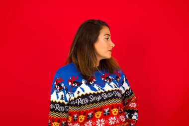 Kırmızı duvara karşı Noel süveteri giyen mutlu, beyaz bir kadının yan görüntüsü.