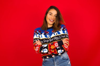 Beyaz kadın kırmızı duvara karşı Noel süveteri giyiyor mutlu gülümsüyor ve kameraya kendinden emin bir şekilde kollarını kavuşturuyor. Pozitif ve kendinden emin kişi.