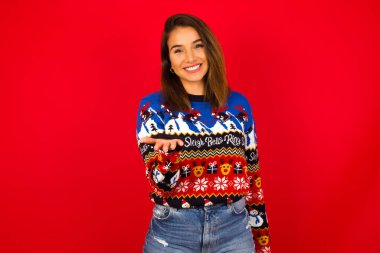 Beyaz kadın, kırmızı duvara karşı Noel kazağı giyip dostça gülümsüyor. Karşılama ve hoş geldin demek için elini ya da elini sıkacak bir şey sunuyor. Başarılı bir iş..