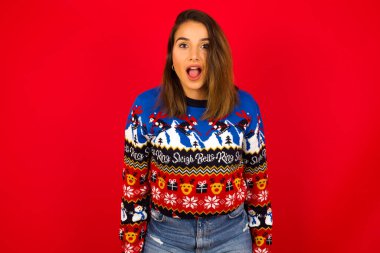 Kafkasyalı bir kadın kırmızı duvara karşı Noel süveteri giyiyor ağzı açık ve şoke olmuş bir şekilde 