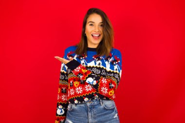 Beyaz kadın kırmızı duvara karşı Noel süveteri giyiyor ve diyor ki: Vay be, ne kadar heyecan verici, hayret verici bir ifade, boş alanda avuç içi ile bir şeyler gösteriyor. Reklam kavramı.