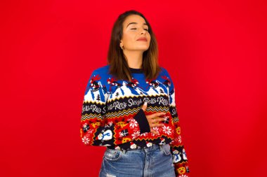 Kırmızı duvara karşı Noel süveteri giyen mutlu, beyaz kadın ellerini karnının üstüne koyuyor, lezzetli bir akşam yemeği yedikten sonra iyi bir ruh hali içinde olması, doyduğunu gösteriyor. Midemde hoş bir his var..