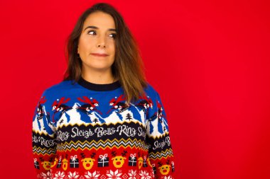 Şaşırmış genç, beyaz bir kadın Noel süveterini kırmızı duvara dayamış, dudakları kıvrılmış ve endişeli görünüyor, önünde korkunç bir şey görüyor..