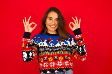 Kırmızı duvara karşı Noel süveteri giyen güzel beyaz bir kadın her iki elinde de işaret işareti olan neşeli bir ifadeye sahip, iyimser bir ifadeye sahip..