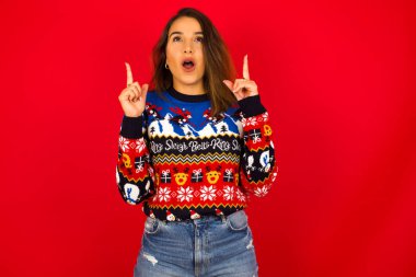 Genç, beyaz bir kadın, kırmızı duvara karşı Noel süveteri giyiyor, şaşırmış ve şaşırmış görünüyor ve parmaklarıyla garip bir şey gösteriyor..