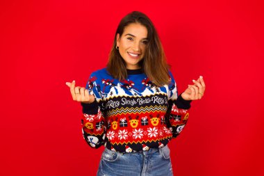 Genç, güzel, beyaz bir kadın Noel süveterini kırmızı duvara dayayıp para hareketi yapıyor..