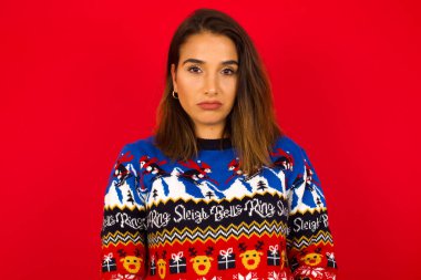 Genç, beyaz bir kadın, kırmızı duvara karşı Noel süveteri giyiyor, gözü yaşlı ve çaresiz bir şekilde ağlıyor, acı ve depresyon çekiyor. Üzgün yüz ifadesi ve duygu konsepti.