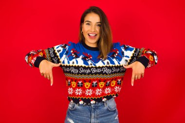 Kırmızı duvara karşı Noel süveteri giyen genç, beyaz bir kadın, olumlu bir ifadeyle, her iki işaret parmağıyla aşağıya işaret ediyor, geniş bir gülümsemeye sahip. Şuraya bakın, lütfen..
