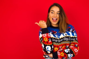 Şaşırmış genç, beyaz bir kadın, kırmızı duvara karşı Noel süveteri giyiyor, şaşırmış bir ifadeyle, gözlerini ve ağzını geniş aralıklarla açıyor, başparmağıyla işaret ediyor, garip bir şey gösteriyor. Reklam kavramı.